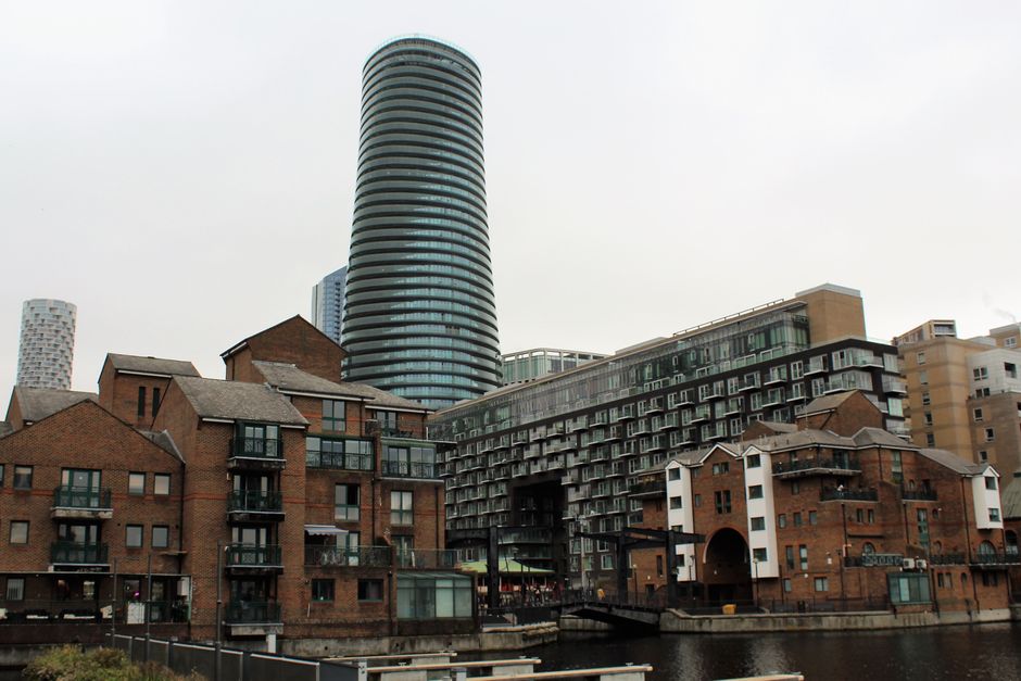 The image shows a waterfront scene in Canary Wharf with modern residential buildings, including a large, dark-colored high-rise tower with a curved design in the background. In the foreground, there are several brick and contemporary style apartment buildings with multiple balconies, some with glass railings, and visible windows. The buildings are situated along a canal or river, and the scene is captured during daytime under overcast weather, with natural light illuminating the area. Near the water, there are outdoor areas with small structures and seating, indicating a mixed-use environment typical of urban residential and leisure spaces within the financial district. This setting reflects a typical context for house and apartment relocations in the Canary Wharf area, where professional removals by companies such as Removal Van Docklands facilitate furniture transport and home relocation logistics.