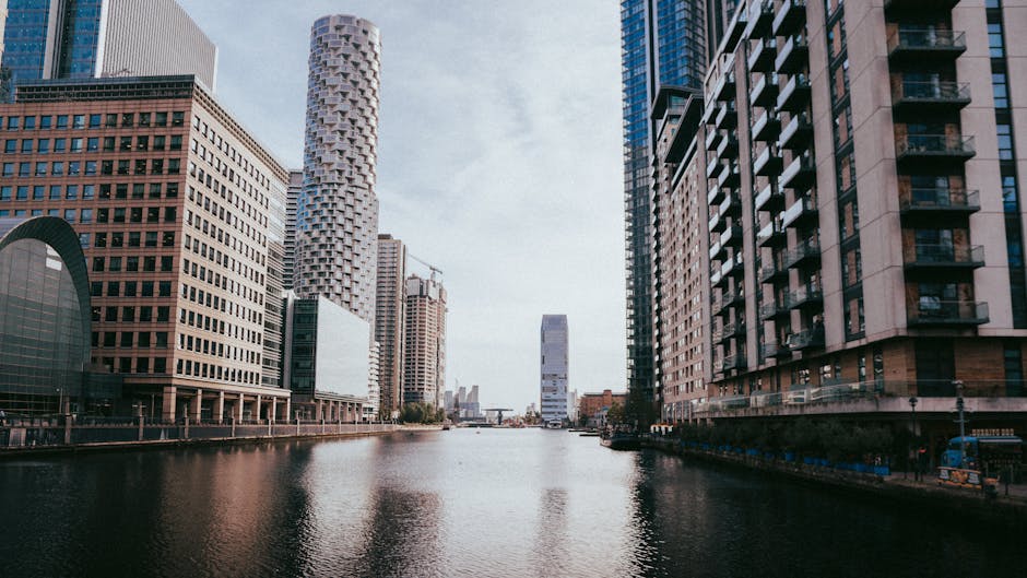 A wide view of the Canary Wharf district in London showing the River Thames with modern high-rise office buildings and residential towers on both sides. The buildings feature a mix of glass and concrete facades, with some structures under construction visible in the background. The river reflects the surrounding skyscrapers, and the sky above is partly cloudy. The scene captures the urban environment where house removals and furniture transport services by Removal Van Docklands are often necessary for residents moving in or out of these tall residential and office towers in E14, Docklands. The image highlights the typical setting for home relocation within a bustling financial district and provides context for the process of packing, loading, and transporting belongings through narrow city streets and alongside waterfront areas.