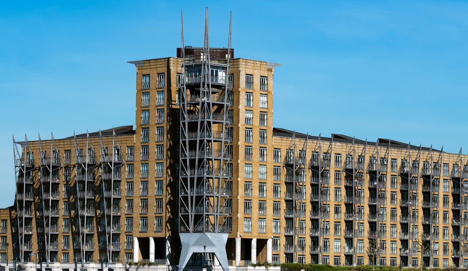 The image shows a waterfront scene in Canary Wharf with modern residential buildings, including a large, dark-colored high-rise tower with a curved design in the background. In the foreground, there are several brick and contemporary style apartment buildings with multiple balconies, some with glass railings, and visible windows. The buildings are situated along a canal or river, and the scene is captured during daytime under overcast weather, with natural light illuminating the area. Near the water, there are outdoor areas with small structures and seating, indicating a mixed-use environment typical of urban residential and leisure spaces within the financial district. This setting reflects a typical context for house and apartment relocations in the Canary Wharf area, where professional removals by companies such as Removal Van Docklands facilitate furniture transport and home relocation logistics.