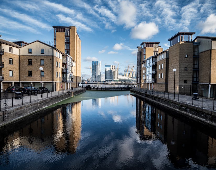 A view of a narrow waterway flanked by modern residential buildings with beige brick exteriors and black metal balconies, situated in the Docklands area. The water is calm, reflecting the partly cloudy blue sky and surrounding structures. On the canal's edge, concrete walkways with black metal fencing are visible, with several parked cars alongside. In the background, a bridge crosses the waterway, leading towards a cluster of high-rise office buildings and cranes, indicating ongoing development. The scene is well-lit with natural daylight, highlighting the urban surroundings. This setting exemplifies the urban environment of the Isle of Dogs, where house removals by companies like Removal Van Docklands often involve navigating narrow streets and tight spaces for transporting furniture and packed boxes in home relocation and furniture transport services.
