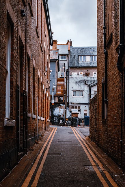 A narrow alleyway between red brick residential buildings on either side, with a double yellow line painted along the pavement. At the far end of the alley, there is a collection of interlinked buildings with various windows, doors, and external staircases, some of which appear to be used for storage or access. The ground is wet, suggesting recent rain, and the scene is lit with natural daylight. Nearby, black waste bins and construction debris are visible, indicating ongoing building or renovation work. This setting reflects the confined spaces often encountered during house removals on the Isle of Dogs, requiring careful planning and equipment for safe furniture transport and home relocation. Natural light highlights the textures of the brickwork and the urban environment where removal services by Removal Van Docklands are frequently called upon to navigate tight, challenging streets.