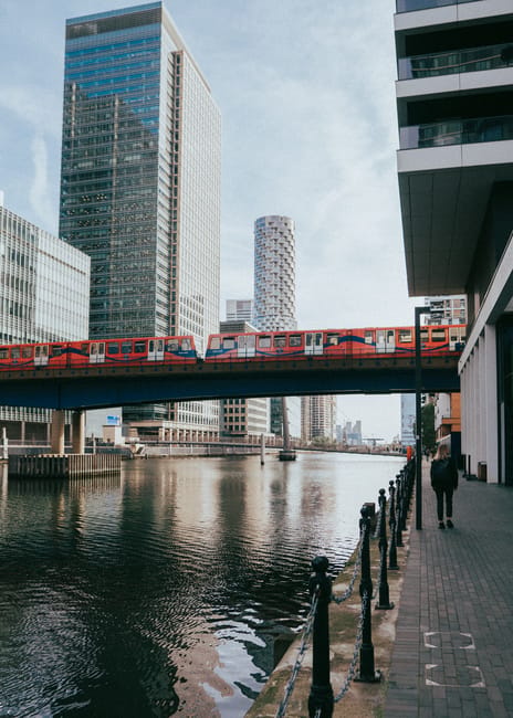 A long, straight outdoor walkway with a metal railing on both sides, leading towards a dock area near the river in an urban setting. The pathway is made of smooth, dark asphalt and is bordered by a ch
